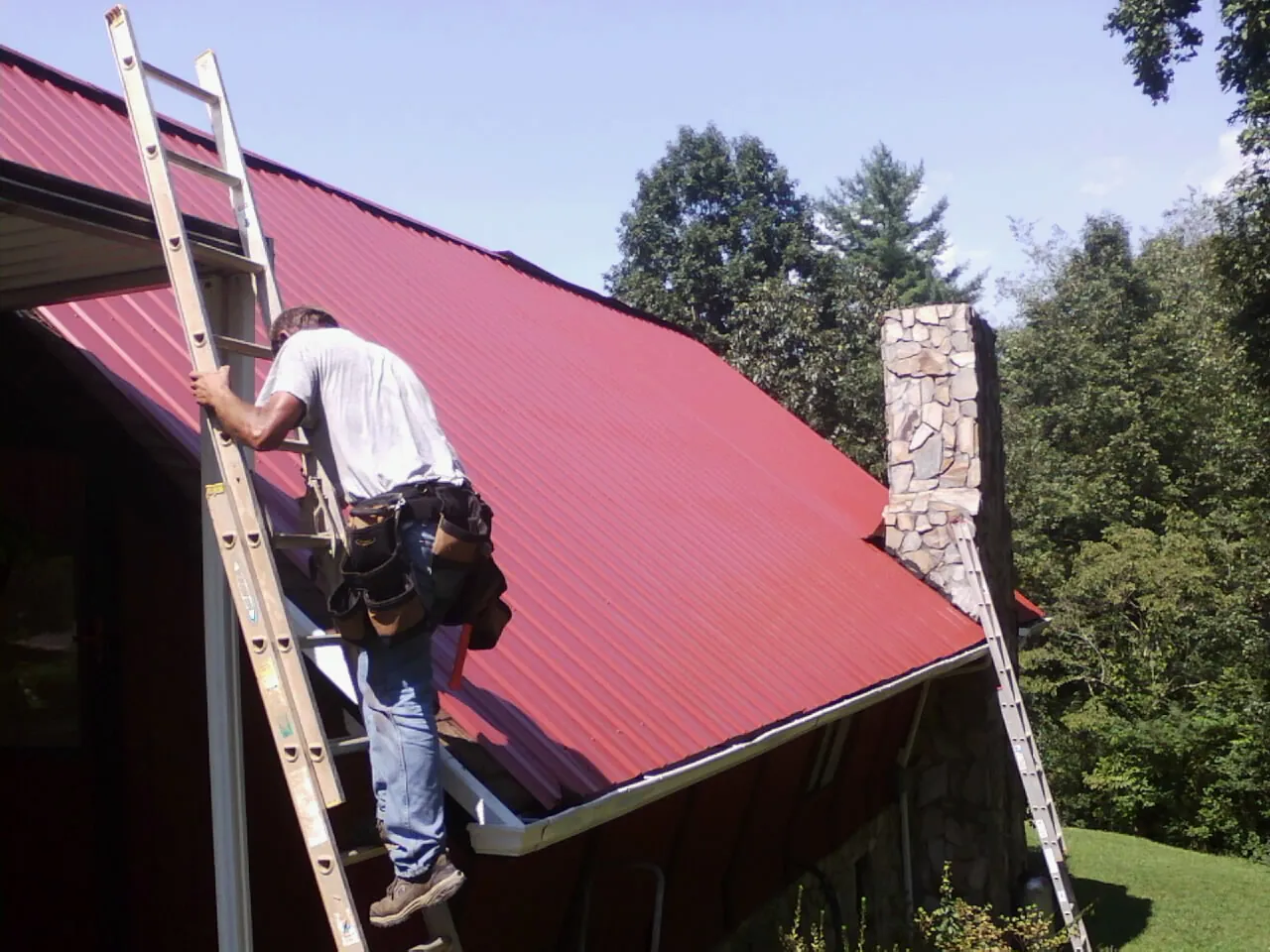 Roofer on a ladder beside a red metal roof and stone chimney — Soffit & Fascia Repair by Roofing Brothers in Unadilla